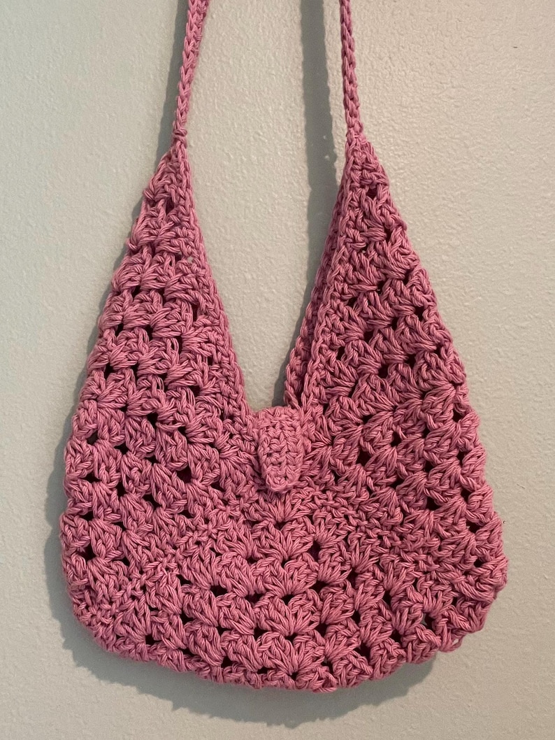 Puede incluir: Bolso de ganchillo rosa hecho a mano con correa para el hombro. El bolso presenta un patr&oacute;n de tejido abierto y texturizado y un peque&ntilde;o cierre de solapa. Cuelga contra una pared azul claro.