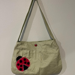 Puede incluir: Un bolso de hombro de color verde claro con una gran aplicación de mariquita roja y negra en la parte delantera. El bolso incluye una correa larga para el hombro, un pequeño cierre de botón de mariquita roja y costuras decorativas negras.