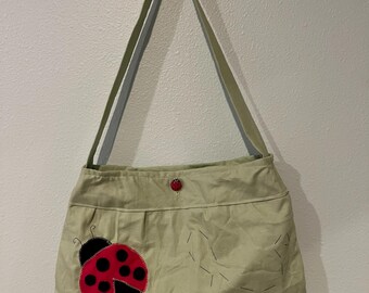 Può includere: Una borsa a tracolla verde chiaro con una grande applicazione a forma di coccinella rossa e nera sul davanti. La borsa presenta una lunga tracolla, una piccola chiusura con bottone a forma di coccinella rossa e motivi decorativi neri.