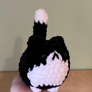May include: A handmade, plush black and white cat. The cat has a round body, small black eyes, and a white face and belly. Its tail is upright, black with a white tip. The cat is made of a soft, textured yarn.