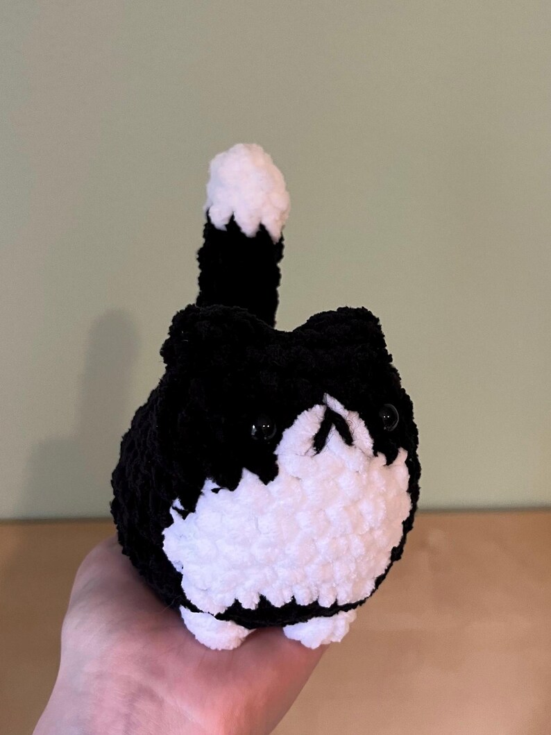 May include: A handmade, plush black and white cat. The cat has a round body, small black eyes, and a white face and belly. Its tail is upright, black with a white tip. The cat is made of a soft, textured yarn.