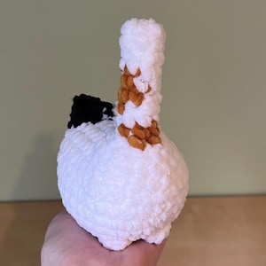 May include: A handmade, white crochet sculpture with a bulbous base and a cylindrical top. The top is accented with brown and tan yarn, and a black section is visible on the side. The sculpture is held in a hand.