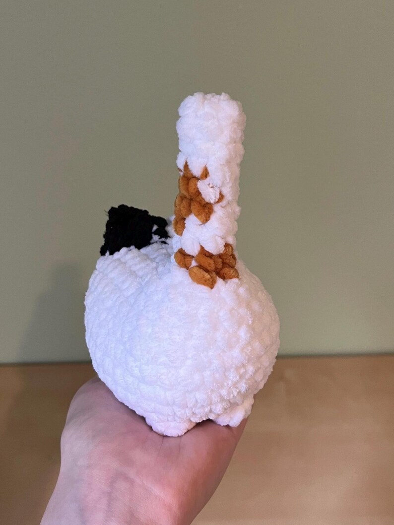 May include: A handmade, white crochet sculpture with a bulbous base and a cylindrical top. The top is accented with brown and tan yarn, and a black section is visible on the side. The sculpture is held in a hand.