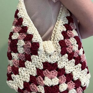 Puede incluir: Bolso de crochet hecho a mano con dise&ntilde;o de granny square. El bolso est&aacute; hecho con hilo crema, burdeos y rosa. La correa y el cierre son de color crema. El bolso es un bolso de hombro.