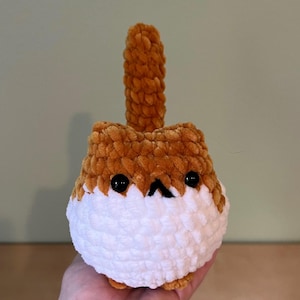 May include: A handmade, plush cat toy in shades of brown and white. The cat has black eyes and a small black nose. The toy is held in a hand, showcasing its soft, textured appearance. The cat has a tail that is standing up.