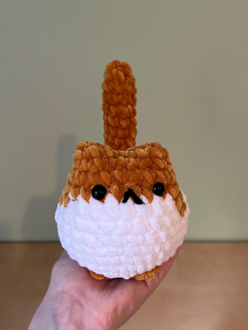 May include: A handmade, plush cat toy in shades of brown and white. The cat has black eyes and a small black nose. The toy is held in a hand, showcasing its soft, textured appearance. The cat has a tail that is standing up.