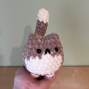 May include: A handmade crochet cat plushie in shades of brown and white. The cat has black eyes and a small, triangular nose. The tail is upright and has a white tip. The plushie is soft and cuddly, perfect for a child's toy or a decorative item.
