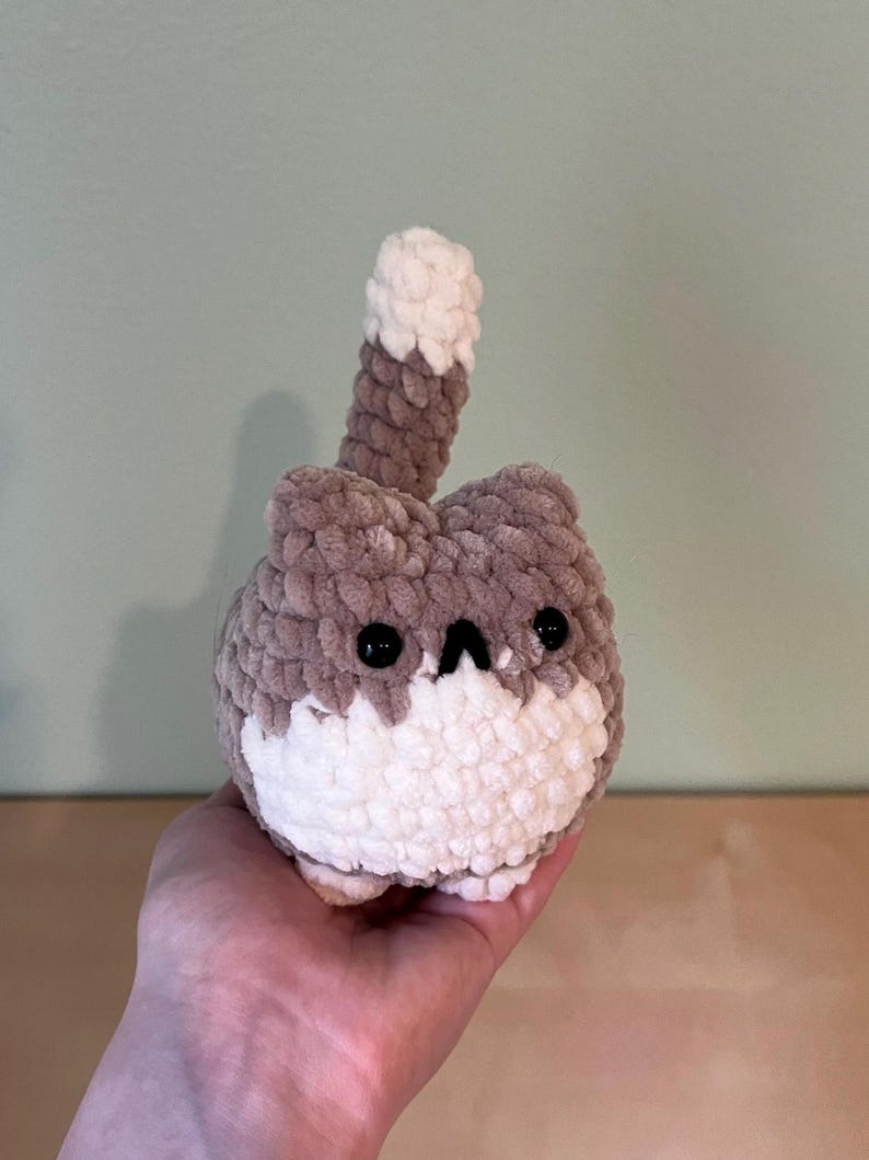 May include: A handmade crochet cat plushie in shades of brown and white. The cat has black eyes and a small, triangular nose. The tail is upright and has a white tip. The plushie is soft and cuddly, perfect for a child's toy or a decorative item.