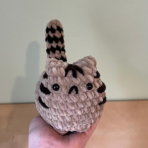 May include: A handmade, plush cat toy in a light brown color with dark brown stripes and a tail. The cat has black button eyes and a small, stitched mouth. The toy is held in a hand.