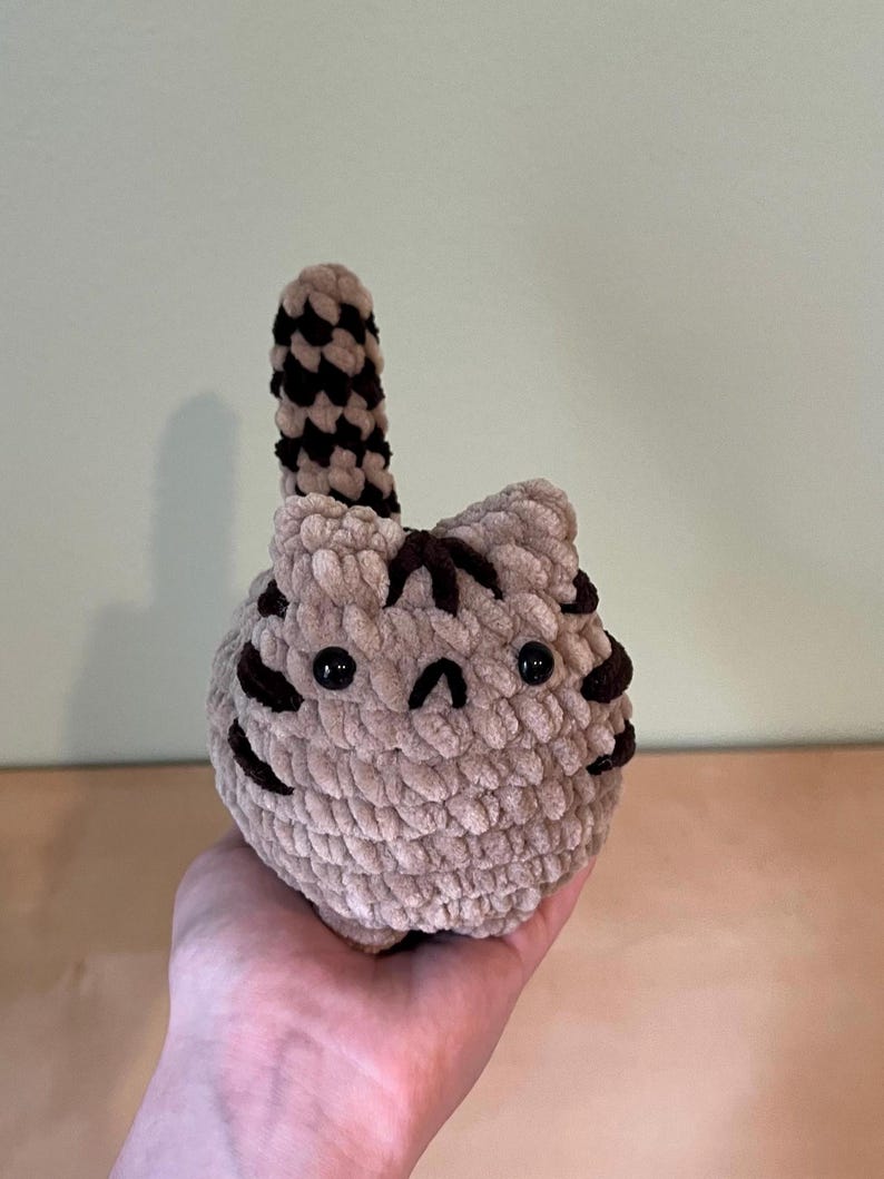 May include: A handmade, plush cat toy in a light brown color with dark brown stripes and a tail. The cat has black button eyes and a small, stitched mouth. The toy is held in a hand.