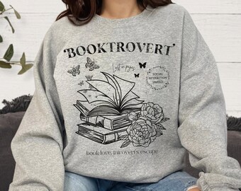 May include: A gray sweatshirt with the text "Booktrovert" and a graphic of a stack of books with butterflies and flowers. The text below the graphic reads "book love, introvert's escape".