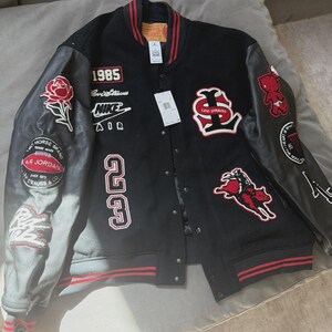 Może przedstawiać: Czarna kurtka typu varsity ze skórzanymi rękawami i licznymi haftowanymi naszywkami, w tym numerem 23, rokiem 1985, brandingiem Nike Air, czerwoną różą, jeźdźcem na byku i logo Levi Strauss, z czerwono-czarnymi prążkowanymi ściągaczami.