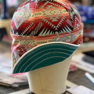 Può includere: Un berretto da saldatura color turchese e bianco con un motivo geometrico in tonalità di rosso, rosa e beige.