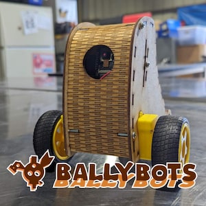 May include: A wooden robot with black wheels and a small camera on the front. The robot is painted brown and has a wood grain pattern. The robot is labeled "BallyBots".