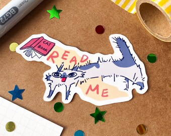 May include: A die-cut sticker featuring a white and blue cat with a wide-eyed expression and tongue sticking out. The cat is positioned next to a pink book labeled CAT BOOK. The text READ ME is written in red letters across the sticker design.