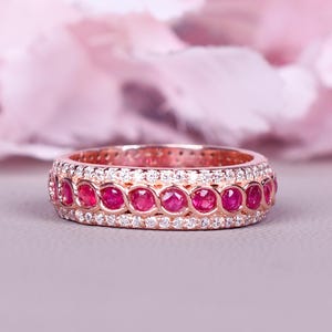 May include: Rose gold ring with a band of oval-cut rubies and small, round diamonds. The ring is set on a light gray surface with a soft pink floral background.