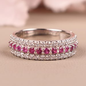 Puede incluir: Un anillo plateado con tres filas de piedras preciosas. Las filas superior e inferior están engastadas con pequeñas piedras transparentes, mientras que la fila central presenta una línea de piedras preciosas de color rosa vibrante. El anillo se encuentra sobre un fondo suave y neutro.