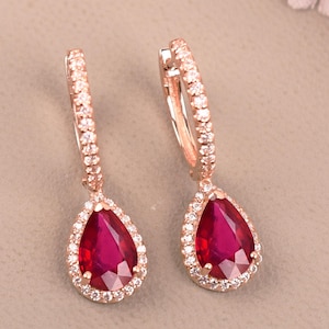 May include: A pair of rose gold earrings featuring a teardrop-shaped, deep red gemstone, encircled by small, clear crystals. The earrings are set against a neutral backdrop, with soft pink floral decorations.