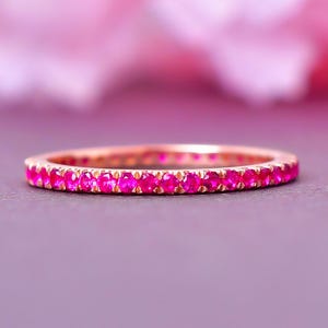 May include: A rose gold eternity ring featuring a continuous line of bright pink gemstones. The ring is set against a soft purple backdrop, with a blurred pink floral element in the background.