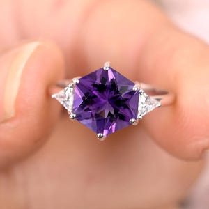 May include: A silver ring featuring a large, square-cut, deep purple gemstone as the centerpiece. Two smaller, triangular white gemstones flank the main stone. The ring is held up against a soft, blurred background.