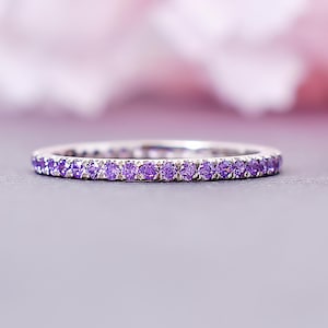 May include: A silver band ring with a continuous row of small, round, purple gemstones. The ring is set against a soft, blurred background of pink and purple hues, creating a delicate and elegant aesthetic.
