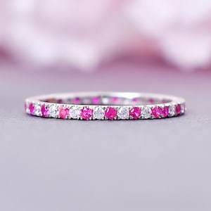 May include: A close-up of a silver ring with alternating pink and clear gemstones. The ring is set against a soft, blurred background with pink and purple hues. The gemstones are round and evenly spaced around the band.