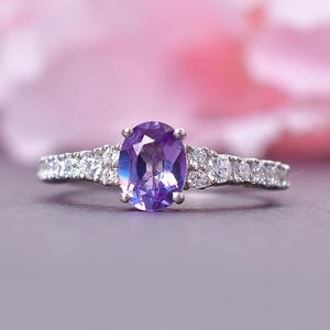 May include: A silver ring featuring a large, oval-cut purple gemstone. The band is embellished with smaller, clear gemstones. The ring is set against a soft pink and grey background, creating a delicate and elegant aesthetic.