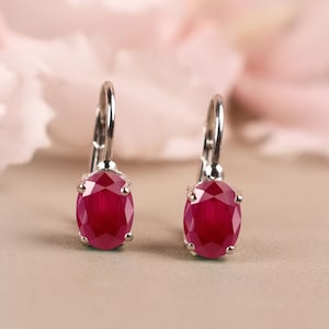May include: A pair of silver leverback earrings, each featuring a vibrant, oval-cut ruby gemstone. The rubies display a deep red hue, set against a soft, neutral background. The earrings are designed for pierced ears.