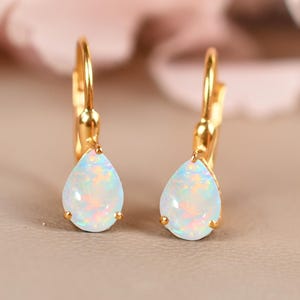 May include: A pair of gold-colored leverback earrings, each featuring a pear-shaped, iridescent opal stone. The earrings are set against a soft, neutral background with blurred pink floral accents.