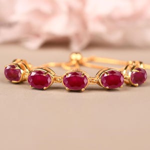 Könnte beinhalten: Ein goldfarbenes Armband mit ovalen, tiefroten Edelsteinen. Die Edelsteine sind in einzelnen Fassungen gefasst und durch eine filigrane Kette verbunden. Das Armband hat einen verstellbaren Schiebeverschluss. Der Hintergrund ist ein weiches, verschwommenes Rosa.
