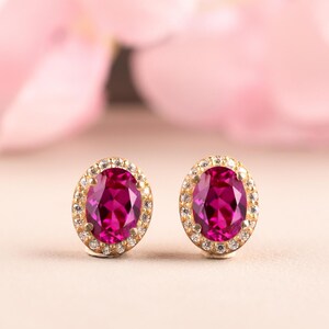 May include: A pair of oval-shaped stud earrings featuring vibrant magenta-colored center stones. Each central gemstone is surrounded by a halo of small, clear, sparkling white stones set in a polished gold-toned metal frame with secure prongs.