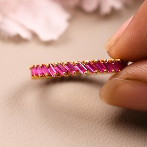 May include: A gold band ring featuring a continuous row of vibrant pink baguette-cut stones set in a channel style. The ring displays a series of rectangular gemstones arranged horizontally around the circumference of the thin metallic band.