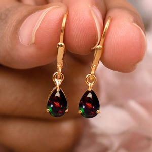 May include: A pair of gold-colored leverback earrings, each featuring a teardrop-shaped black opal with red and green flashes. The earrings are held up, showcasing their design and color.