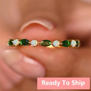 May include: A gold ring with alternating oval green gemstones and round white gemstones. The ring is held up by a person. The text "Ready To Ship" is on a pink rectangle.