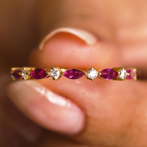 May include: A gold band ring featuring alternating marquise-cut ruby gemstones and round, clear diamonds. The ring is held up by a person's fingers, showcasing the ring's delicate design and vibrant colors.