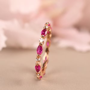 May include: A gold ring featuring alternating marquise-cut ruby and round diamond gemstones. The ring has a delicate design with the gemstones set closely together, creating a continuous pattern around the band. The ring is set against a soft, blurred background.