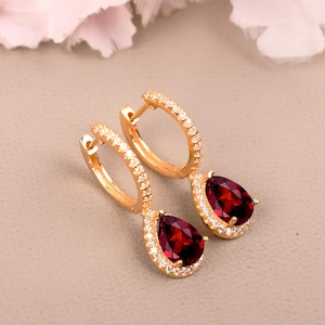 May include: A pair of gold hoop earrings featuring a teardrop-shaped deep red gemstone pendant. Each hoop is encrusted with small clear stones along the front, and the teardrop gemstone is surrounded by a halo of tiny clear stones in a gold setting.