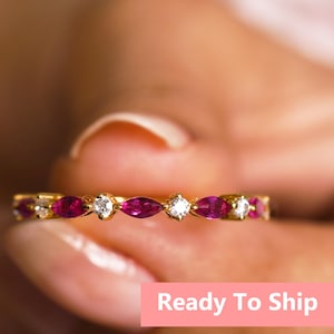 以下が含まれることがあります： ゴールドのリング。マーキス型のルビーとラウンドダイヤモンドが交互に並んでいます。リングは手で持たれており、ピンク色の長方形に白い文字で「Ready To Ship」と書かれています。
