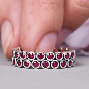 May include: A silver ring with two rows of vibrant red gemstones. The ring is held between two fingers against a soft gray background, with a blurred pink floral element in the background. The gemstones are round and evenly spaced.