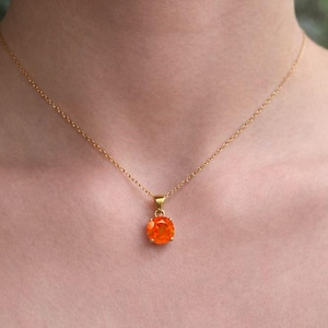 Puede incluir: Un collar dorado con un colgante redondo de piedra preciosa naranja. El colgante cuelga de una cadena fina. El collar se lleva alrededor del cuello, creando un aspecto sencillo pero elegante. La piedra es de color naranja vivo.