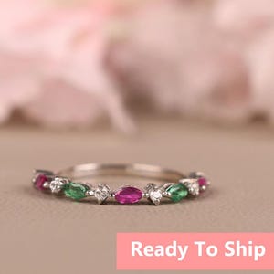 Op de afbeelding: Een zilveren ring met afwisselende robijn, smaragd en diamant edelstenen. De ring is geplaatst op een neutrale achtergrond met een zachte roze en witte wazige achtergrond. De tekst "Ready To Ship" staat in een roze rechthoek.