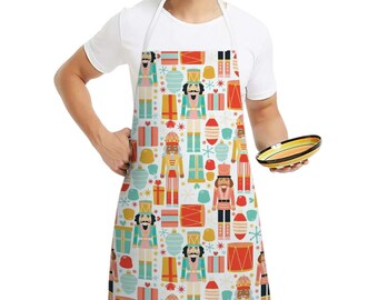 May include: A white apron with a colorful Christmas-themed pattern featuring nutcrackers, gifts, ornaments, and stars. The apron has a neck loop and waist ties. The design includes red, blue, yellow, and green elements.