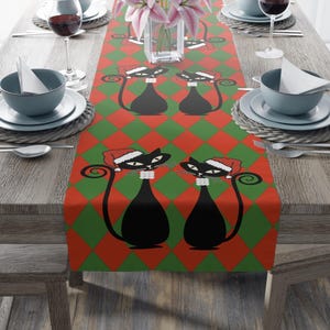 May include: A festive table runner featuring a repeating pattern of black cat illustrations wearing Santa hats, set against a red and green diamond background. The runner is placed on a wooden table, with place settings and glassware visible.