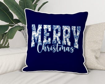 May include: A square, navy blue decorative pillow with the words "MERRY Christmas" in white, floral-patterned letters. The pillow has a white border and is placed on a white sofa.
