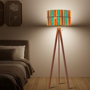 Puede incluir: Una lámpara de pie de estilo retro con un trípode de madera. La pantalla tiene un patrón geométrico en naranja, turquesa y marrón. Un diseño de estrella está centrado en la pantalla. La lámpara es una adición elegante a cualquier habitación.