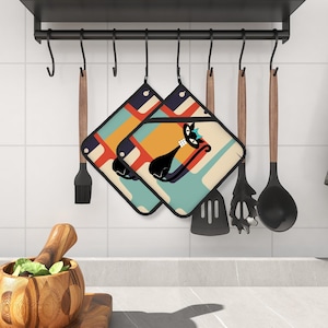 Puede incluir: Un juego de dos agarraderas de olla coloridas con un diseño de gato negro. Las agarraderas de olla tienen un estilo retro, moderno de mediados de siglo con formas geométricas y colores brillantes.