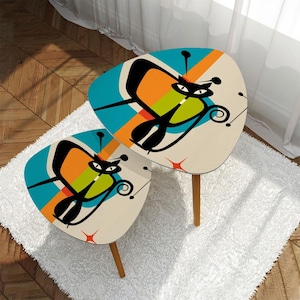 May include: Two retro-style side tables with a mid-century modern design. Each table features a cream-colored surface with a stylized black cat graphic, set against a backdrop of teal, orange, and green stripes. The tables have wooden legs.