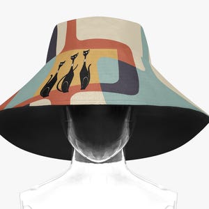 Könnte beinhalten: Ein breitkrempiger Sonnenhut mit einem geometrischen Muster in Schwarz, Weiß und Blau. Drei schwarze Katzen sind auf der Vorderseite des Hutes silhouettiert.