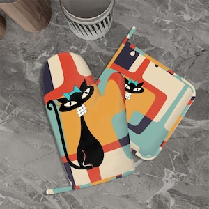 May include: A retro-style oven mitt and pot holder set. The mitt and pot holder feature a black cat design with a blue bow, set against a colorful geometric pattern of orange, red, and teal. The mitt has a long, curved handle.