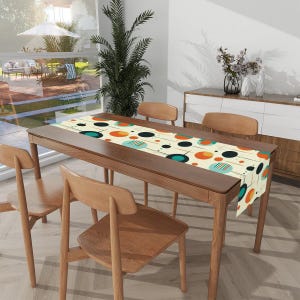 Puede incluir: Un camino de mesa beige con un patrón geométrico de círculos naranja, azul verdoso y negros sobre un fondo blanco. El camino de mesa está colocado sobre una mesa de madera con cuatro sillas.